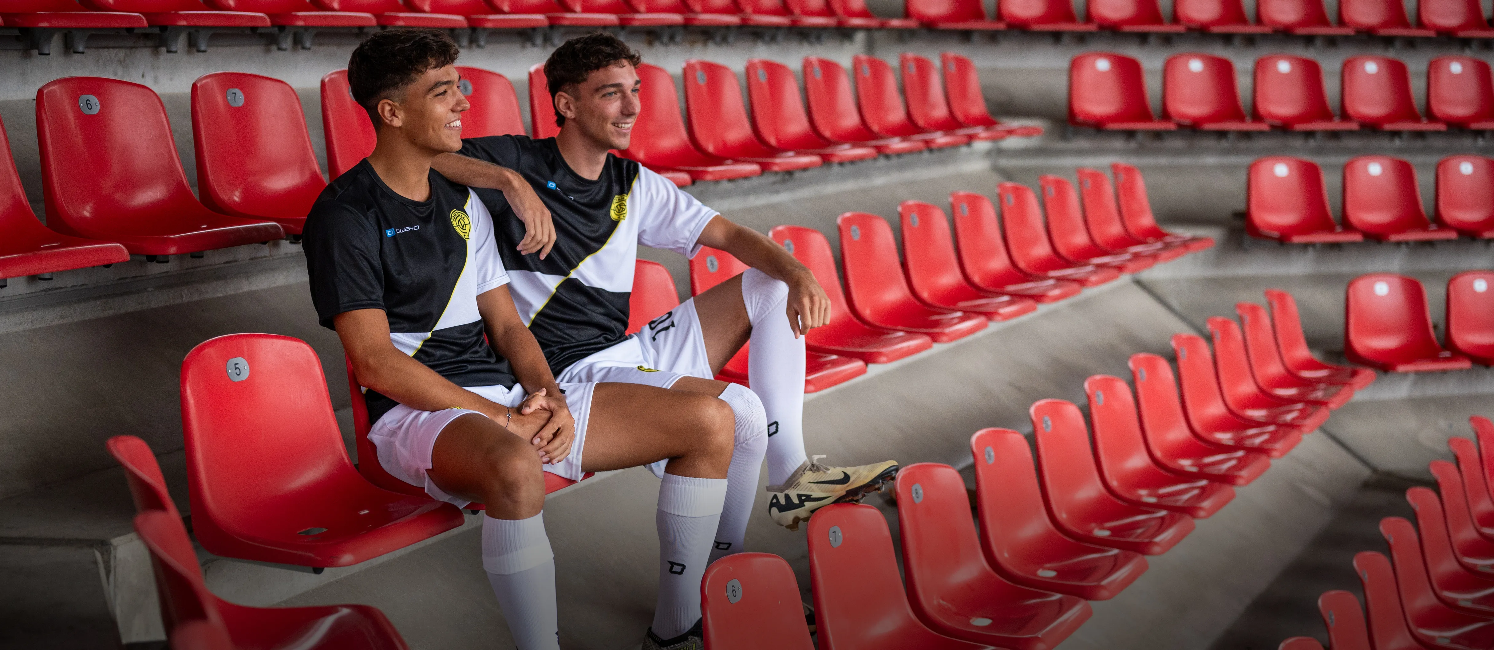 To unge menn i sporty fotballklær sitter avslappet på røde stadionseter.