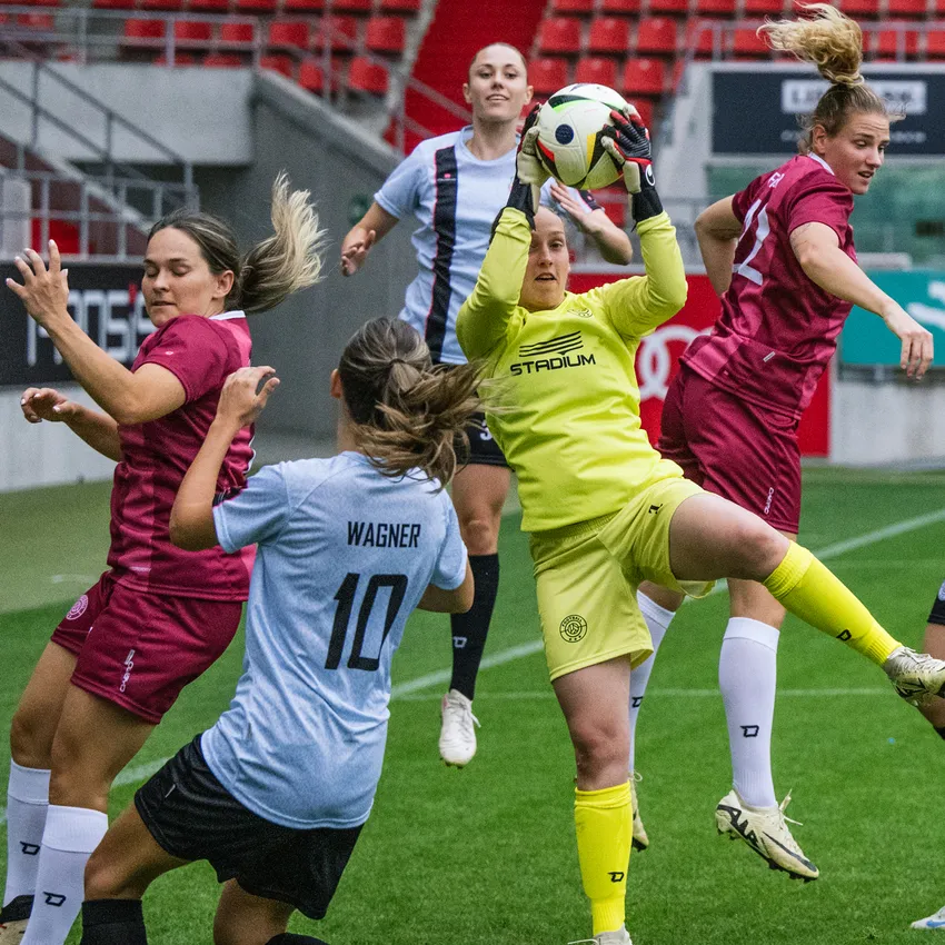 En målvakt i gul sportsklær fanger ballen mens spillere i forskjellige drakter konkurrerer på fotballbanen.