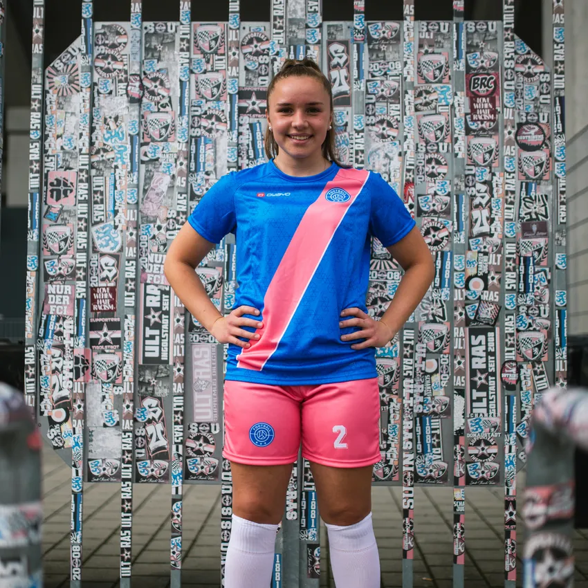 En ung jente i en blå fotballdrakt med en rosa stripe og rosa shorts står foran en vegg dekket med klistremerker.