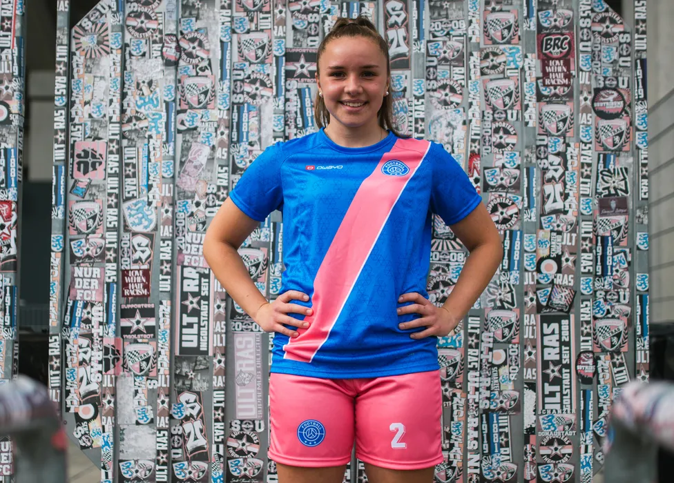 En ung idrettsutøver poserer i en blå fotballdrakt med en rosa stripe og rosa shorts foran en fargerik vegg dekket med klistremerker.