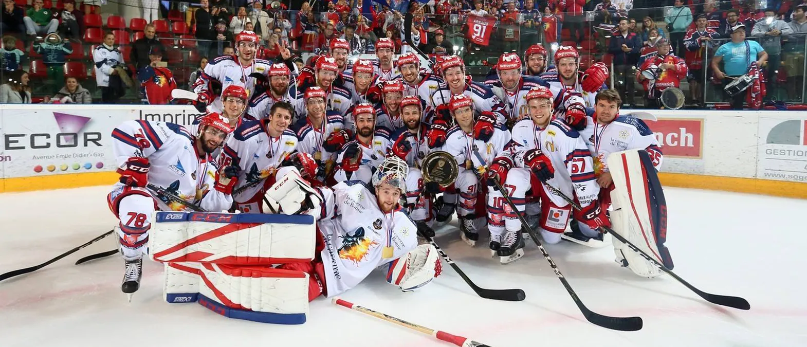 Et ishockeylag i hvite drakter med røde og blå detaljer poserer på isen, omgitt av fans, mens spillerne bærer medaljer og holder en trofé.