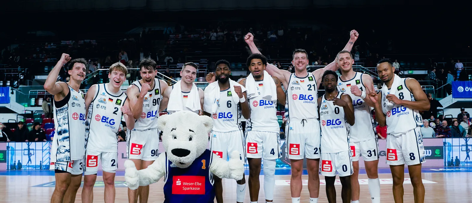 Basketballspillere i hvite trøyer med BLG Logistics-logoet feirer på banen, omgitt av et publikum, med en isbjørn-maskot i forgrunnen.