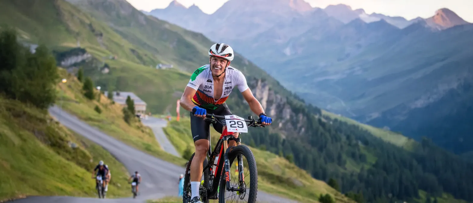 En syklist i en hvit, grønn og oransje trøye med nummer 29 sykler på en fjellvei i et fjellaktig landskap.