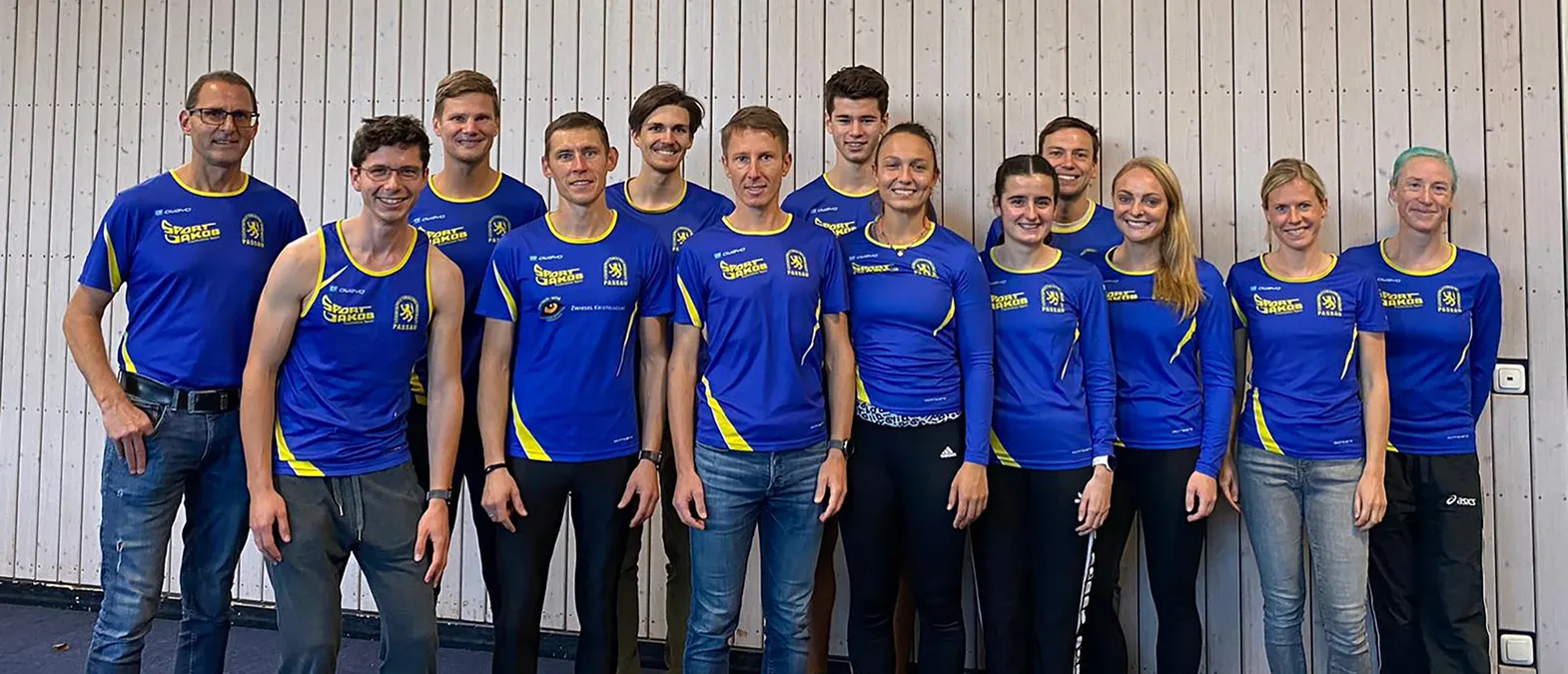 En gruppe menn og kvinner i blå og gule sportstrøyer med logoen 'Sport Jakob Passau' poserer foran en lys trevegg.