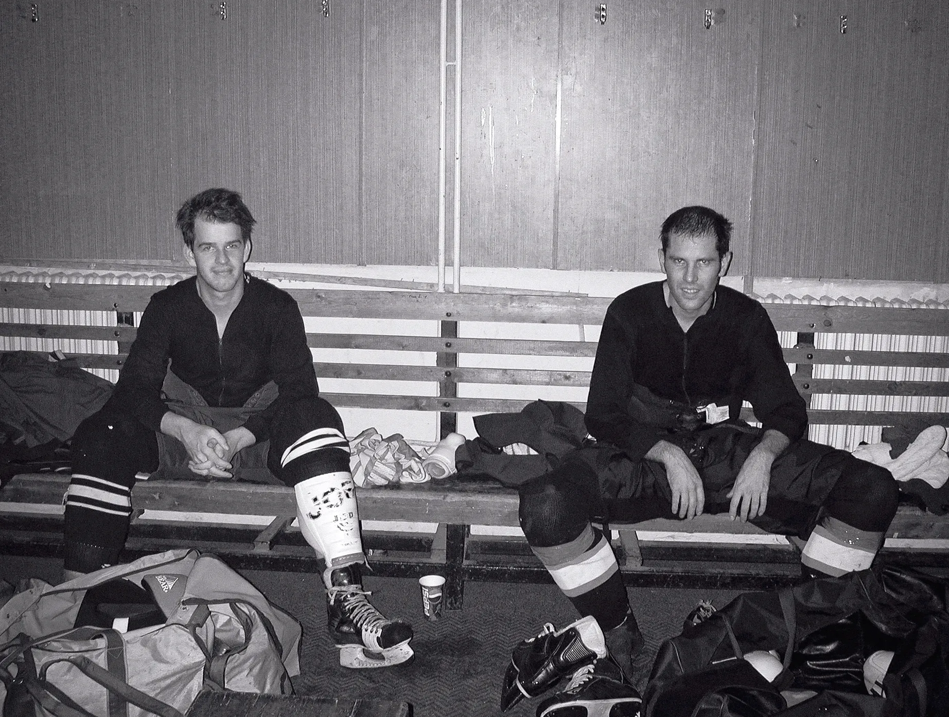 To ishockeyspillere i full utstyr sitter på en trebænk i et omkledd rom, omgitt av sportstasker og skøyter.