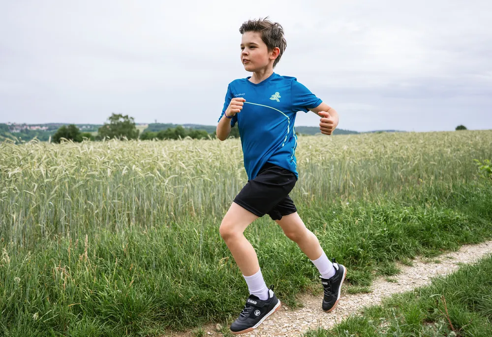 En gutt i en blå sportskjorte og svarte shorts løper langs en sti gjennom et felt.