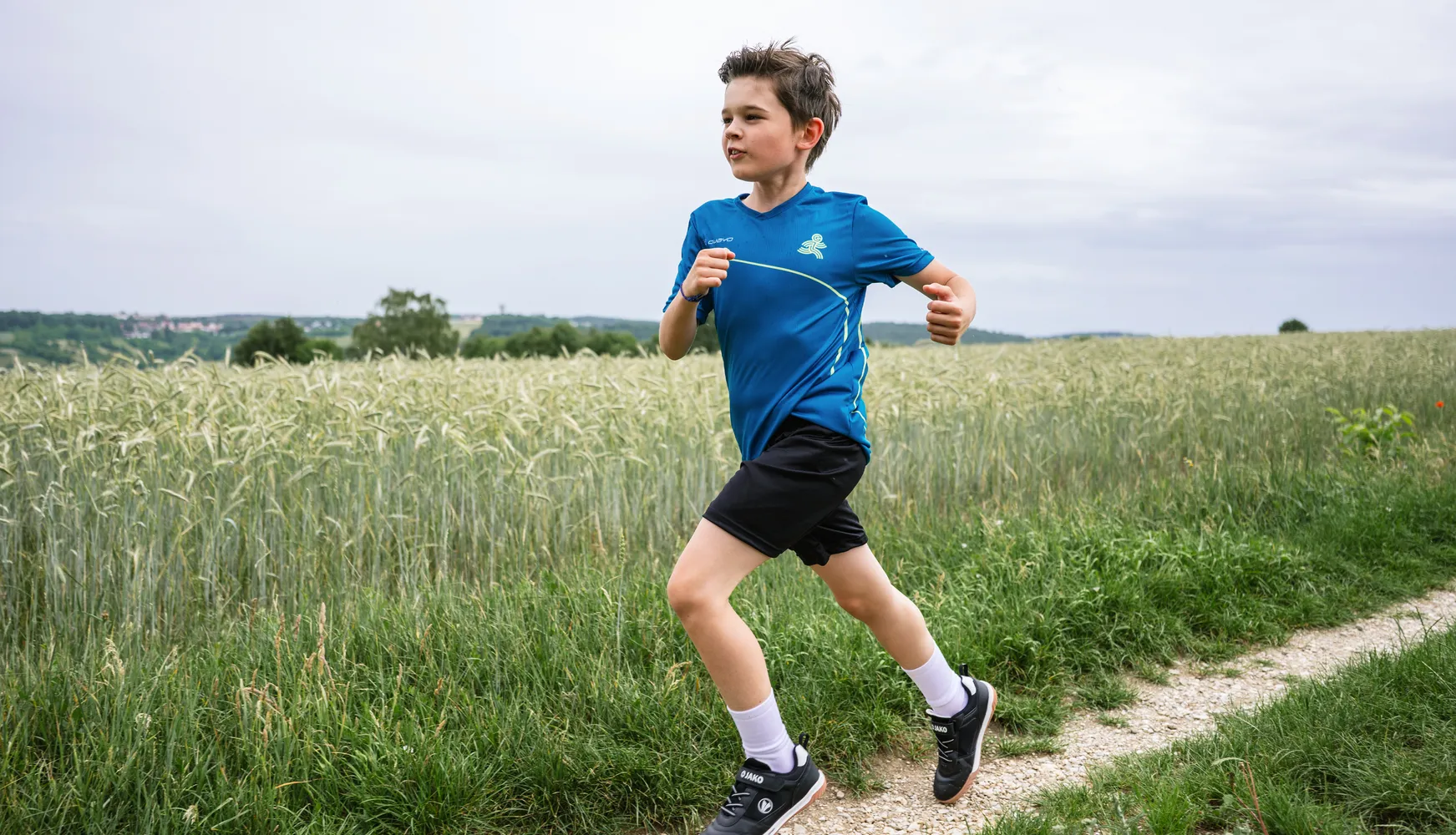 En gutt løper langs en sti gjennom et felt, iført en blå sportstrøye og svarte shorts.