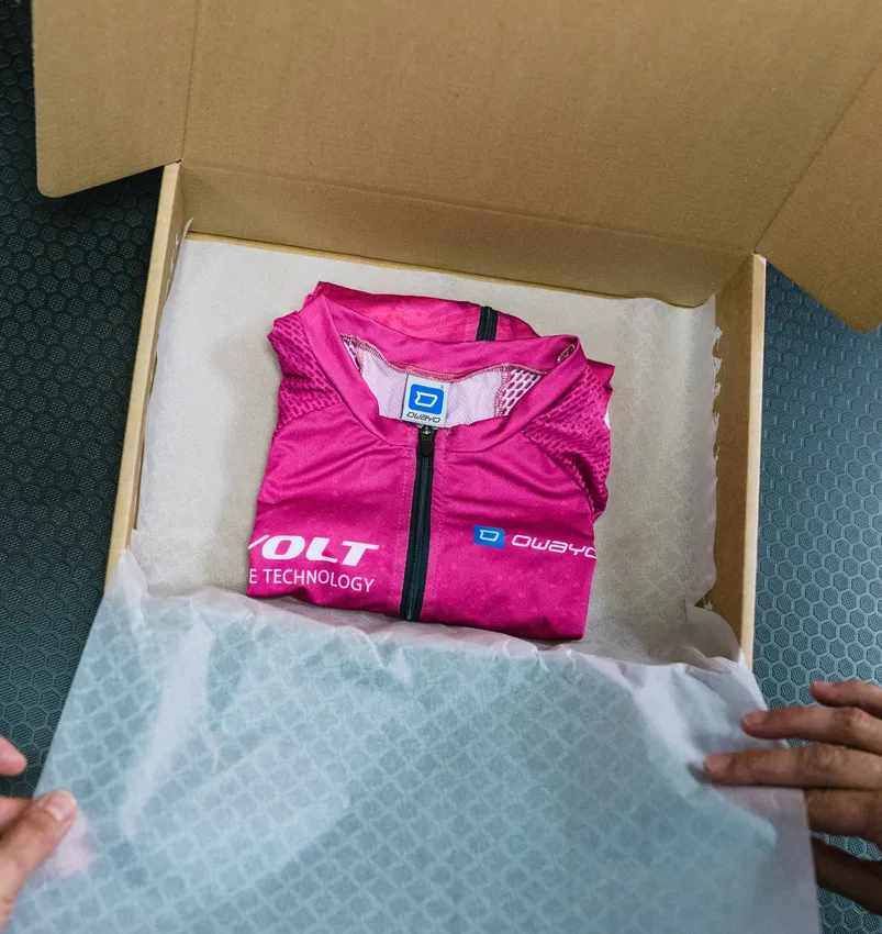 En foldet, rosa, tilpasset sportstøy fra owayo ligger i en åpen pappeske på hvitt papir, holdt av hender.