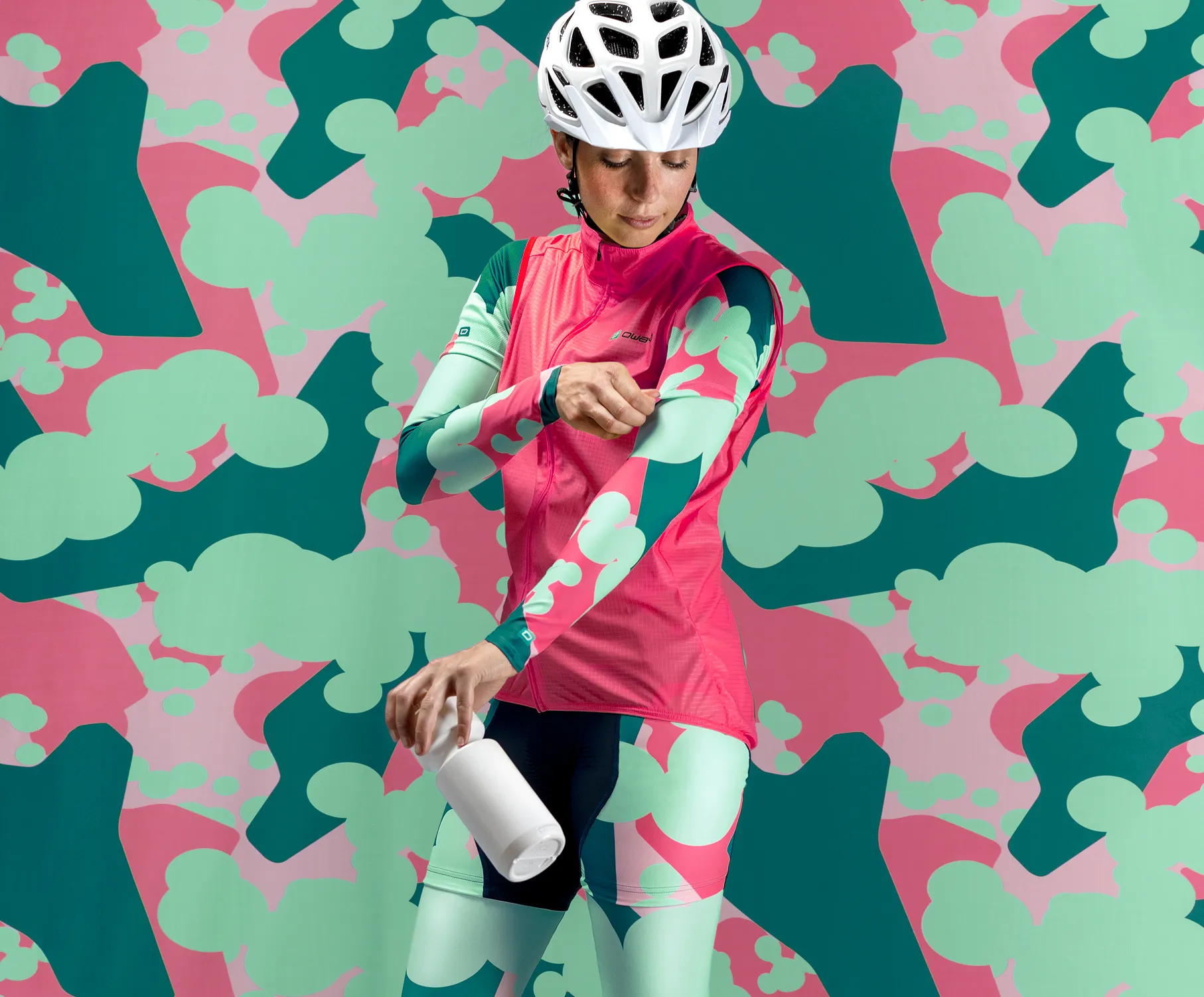 En syklist i tilpasset rosa-grønn sportsutstyr med et abstrakt mønster, som justerer armene sine mens hun holder en hvit flaske, foran en fargerik bakgrunn med samme mønster.