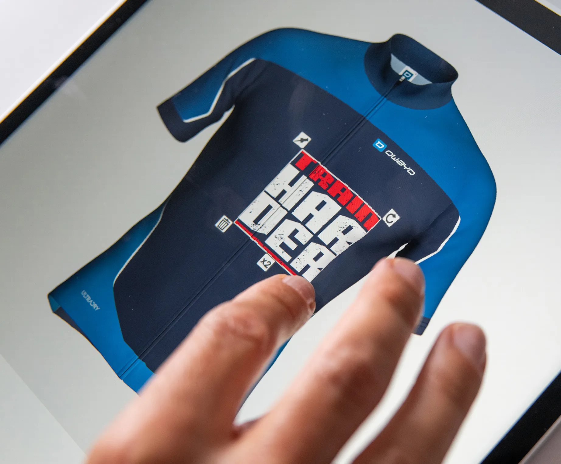 En hånd berører en skjerm som viser et tilpasset mørkeblått og blått sportskjole med teksten 'TRAIN HARDER' og owayo-logoen.