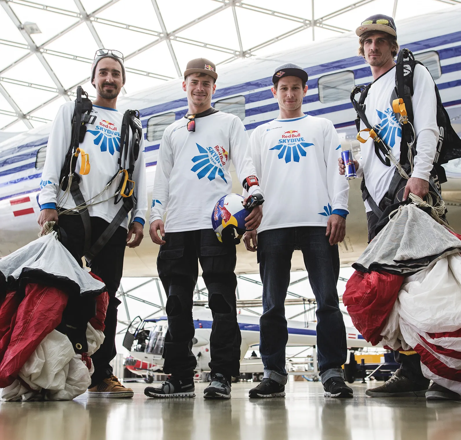 Fire menn i hvite, lange-ermete sportskjorter med Red Bull Skydive Team-logoet står i en hangar foran et fly.