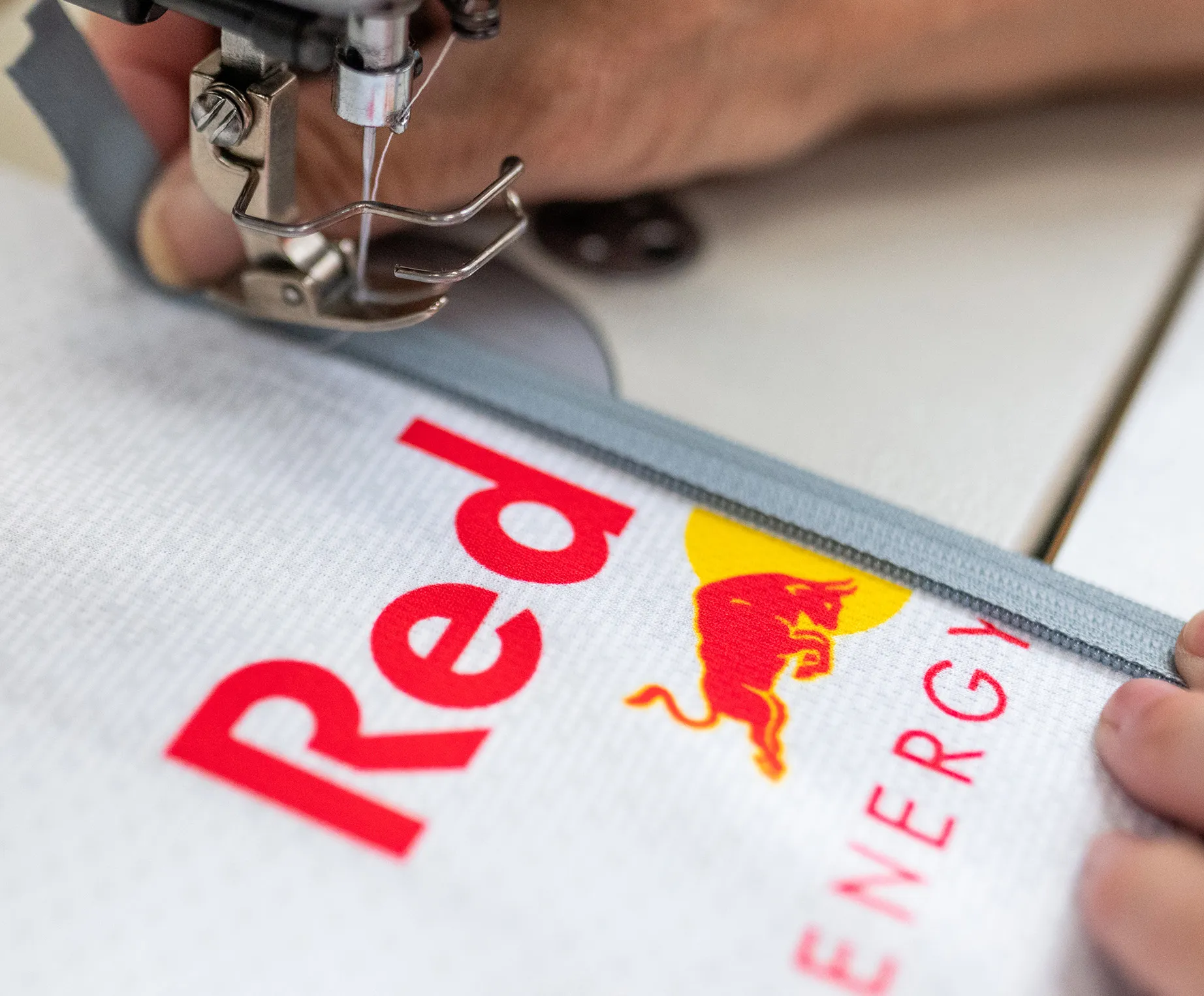 En syemaskin syr en grå glidelås på et hvitt sportstøy med Red Bull-logo.