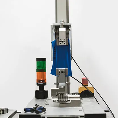 Et blått stoff blir testet i en maskin på et arbeidsbord, ved siden av et signallys og verktøy.