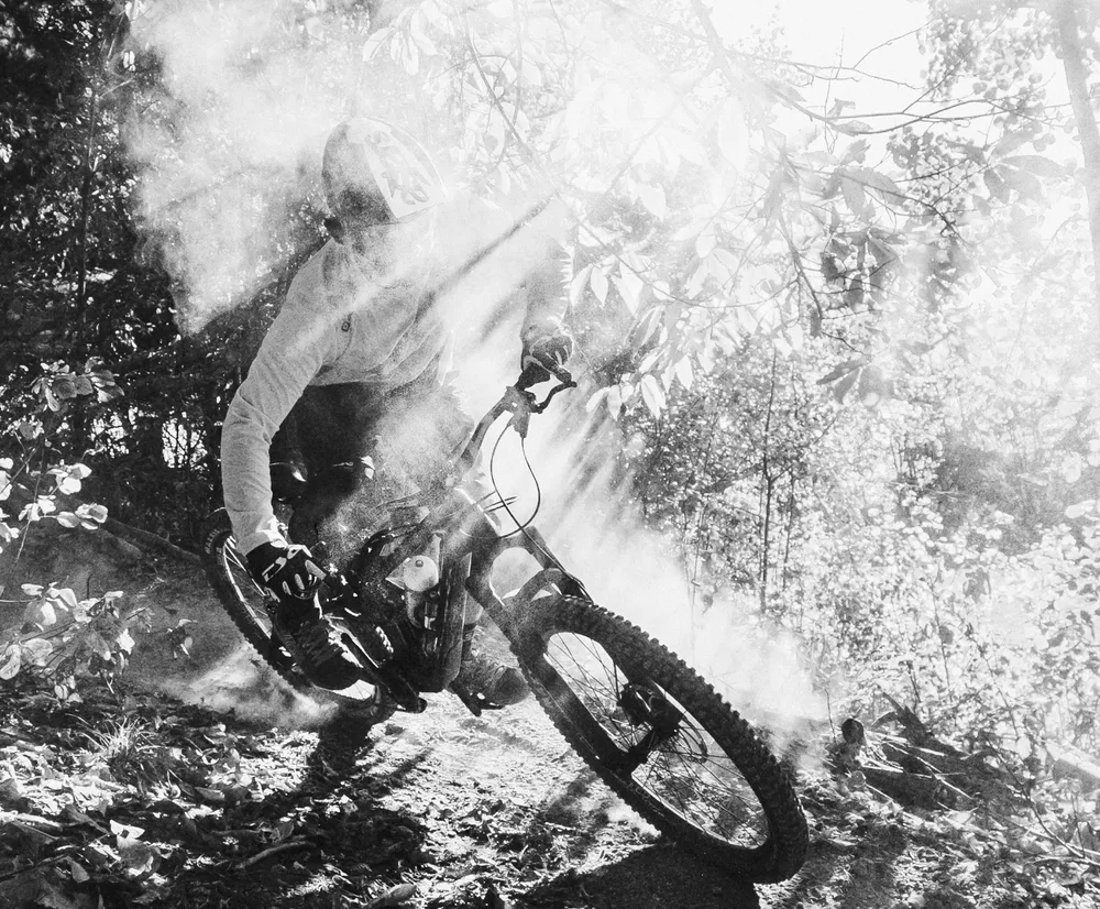 En mountainbiker i bevegelse på en skogsti, omgitt av støv og lysstråler, bærer en langærmet topp fra owayo.