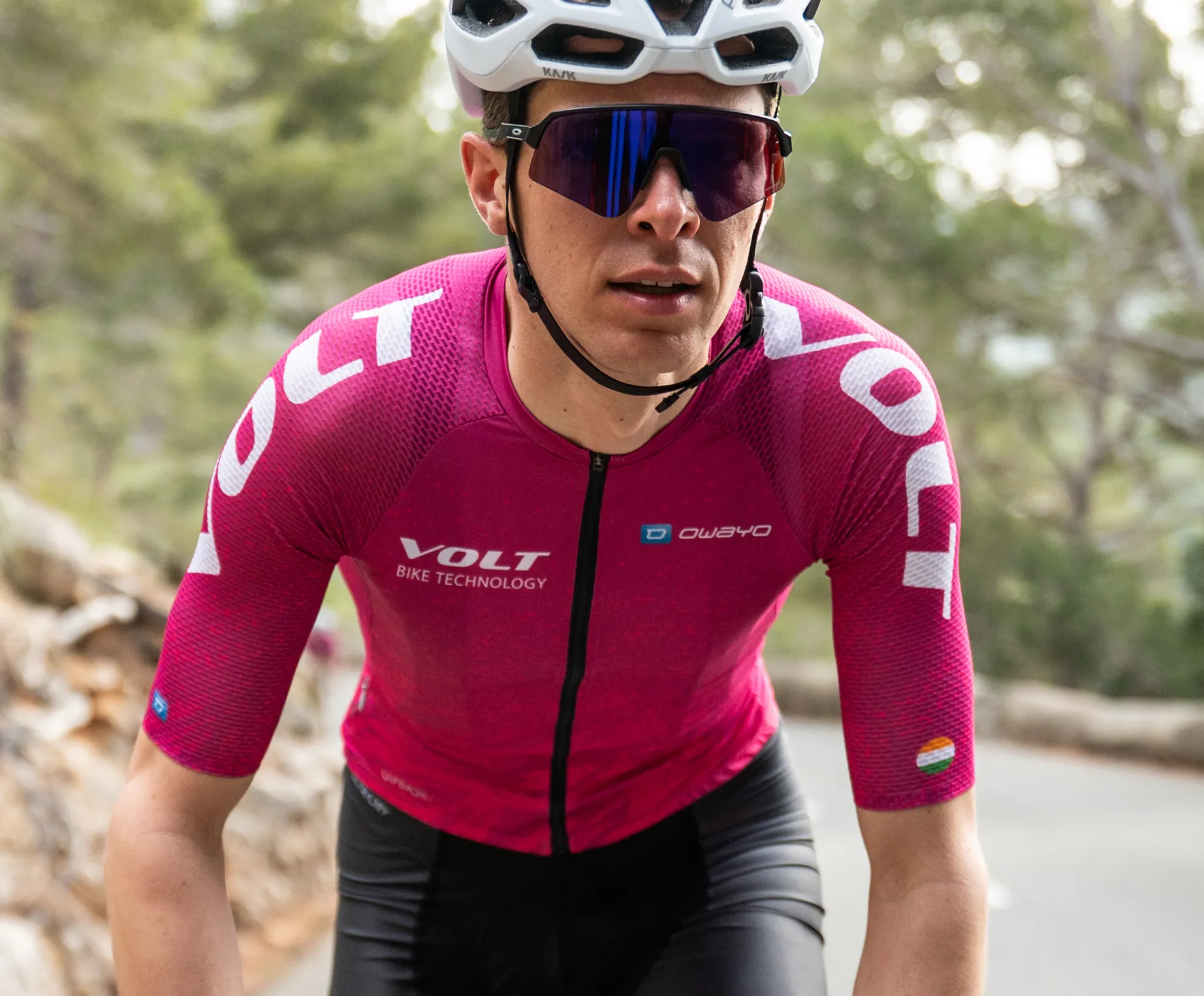 En syklist i en rosa, korte ærmet sykkeltrøye med logoen 'VOLT BIKE TECHNOLOGY' og 'owayo'-logoen på brystet, som sykler på en vei i et skogområde.