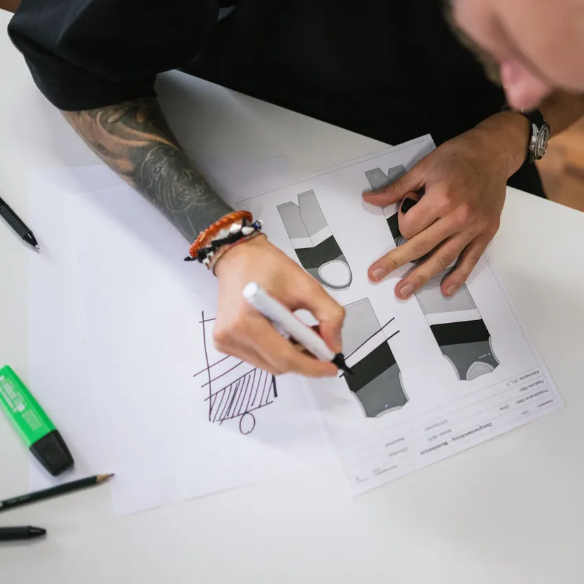 En person skisserer design for sportswear på papir, omgitt av penner og markører.