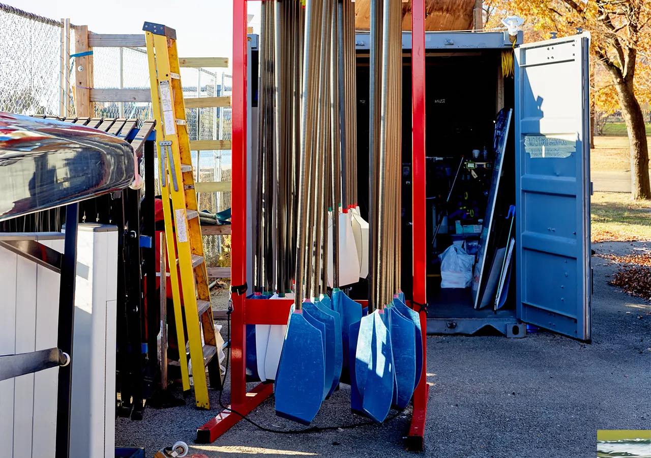 Et bilde av et båtlagringsområde med årer i forskjellige farger, en gul stige og en åpen container i bakgrunnen.