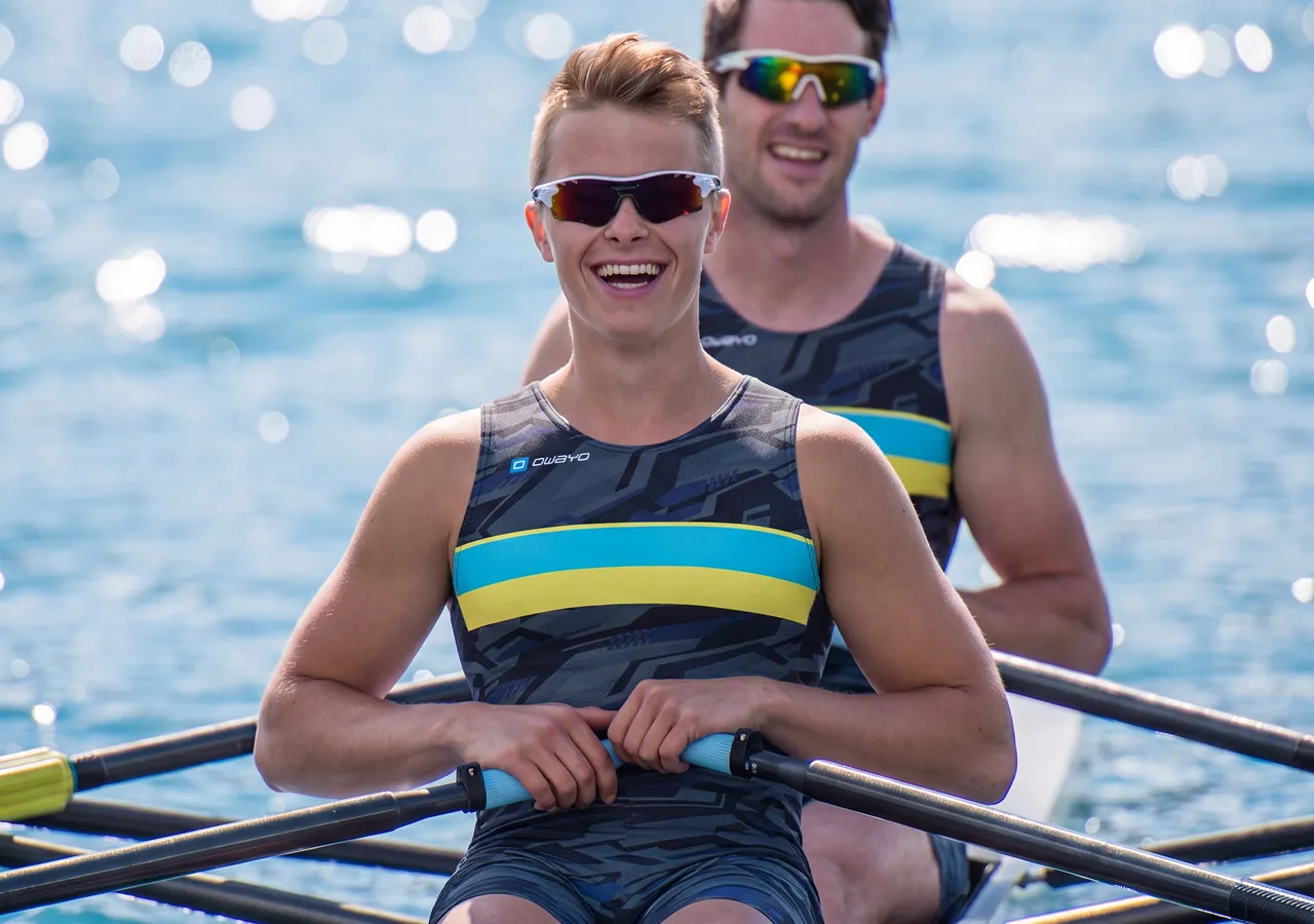 To utøvere i stilige sportsklær sitter i en robåt og smiler til kameraet mens de padler på glitrende vann.