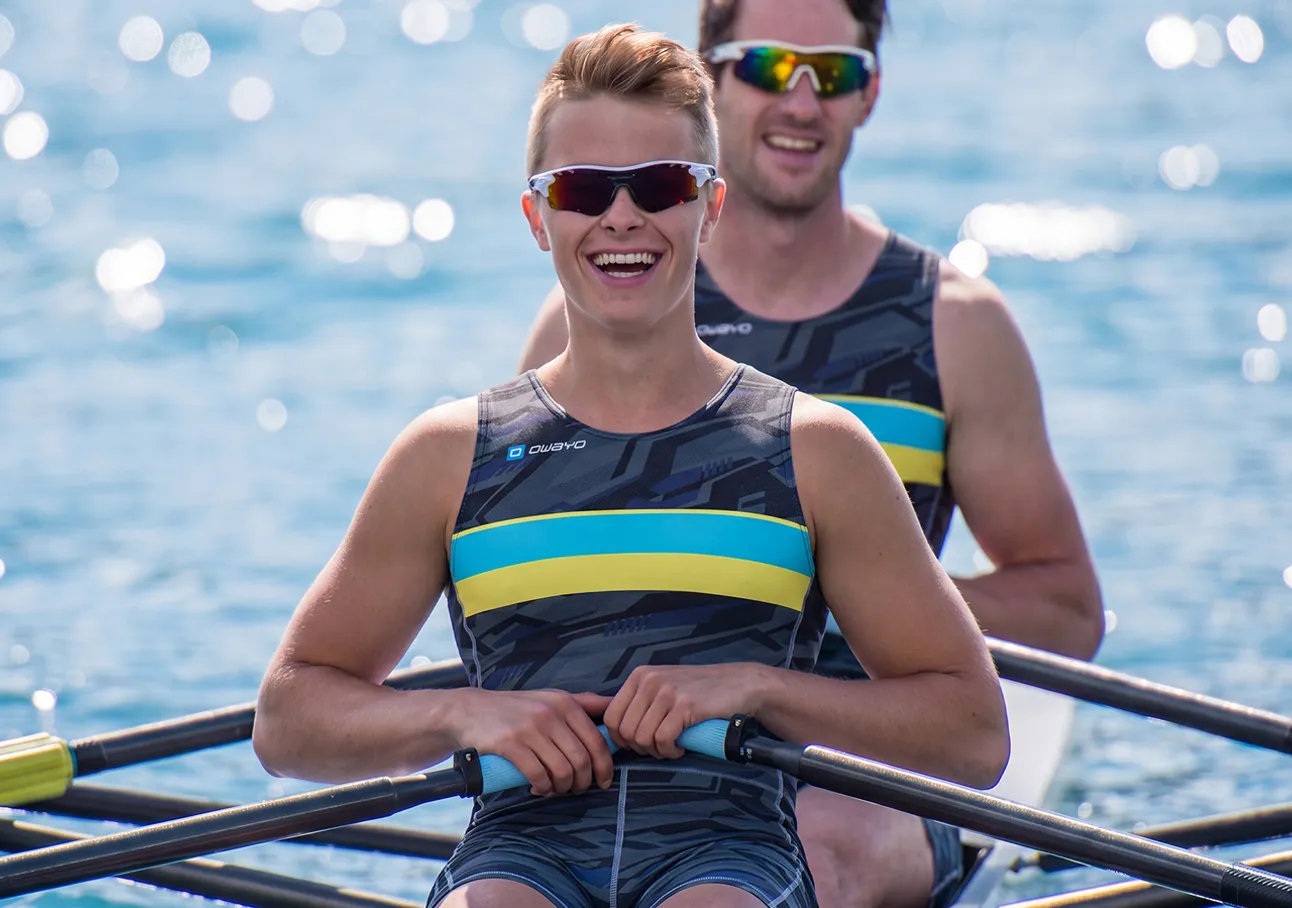 To glade roere i atletiske drakter med blå og gule striper som sitter i en båt på glitrende vann.