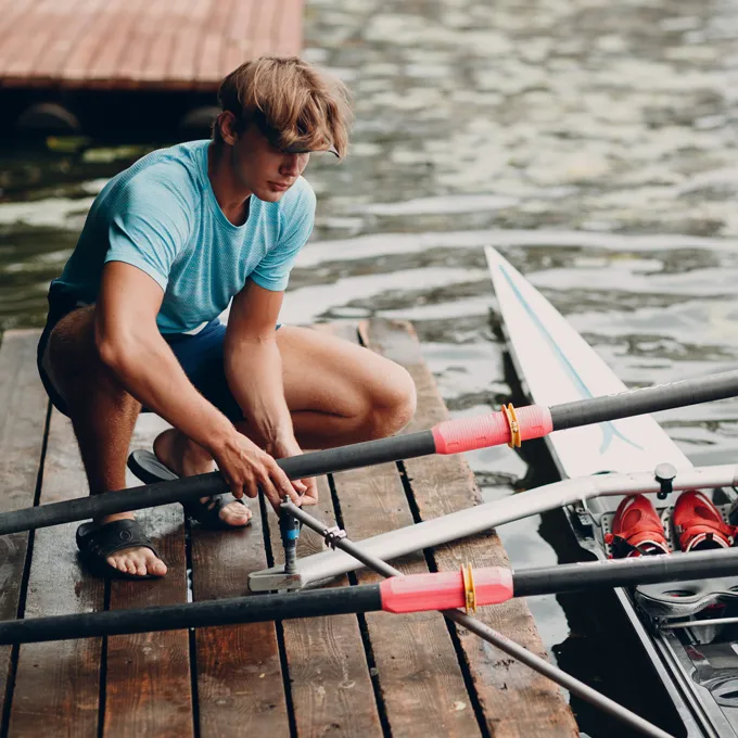 En ung mann i en blå sportskjorte reparerer årer på en brygge ved vannet.