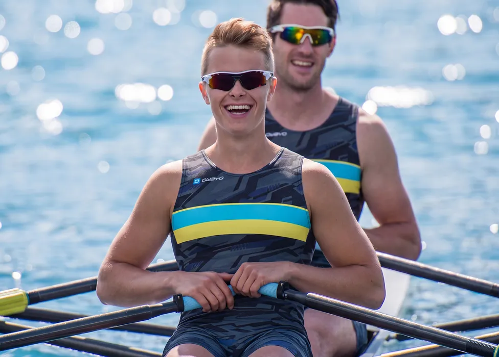 To roere i sporty mønstrede drakter med blå og gule striper, smilende på en vannflate.