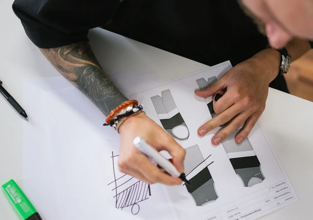 En person med et tatovering på armen tegner med en hvit penn på et ark papir med flere designer av sportstøy.