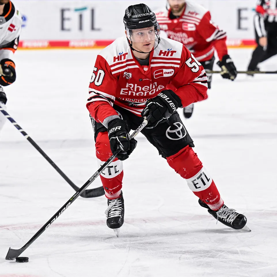 En ishockeyspiller i en rød trøye med nummer 50 og teksten 'Rhein Energie' på isen, som kontrollerer en puck med sin stok.