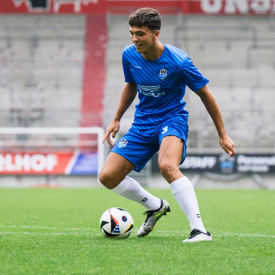 En ung fotballspiller i en blå trøye og blå bukser med et logo på brystet og nummer 3 på bukserne dribbler en fotball på et grønt gressfelt.