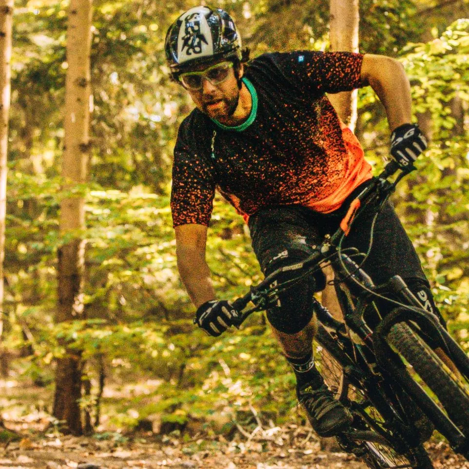 En mountainbiker i en svart, rød prikket trøye og svarte bukser sykler gjennom en skog.