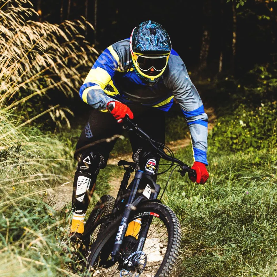 En mountainbiker i en fargerik, langærmet trøye med blå, gul og grå detaljer sykler på en skogsti.