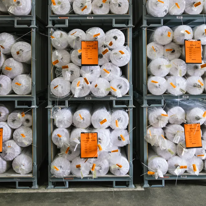 Et lager med hyller fylt med store ruller med hvit stoff pakket i plast og merket med oransje etiketter.