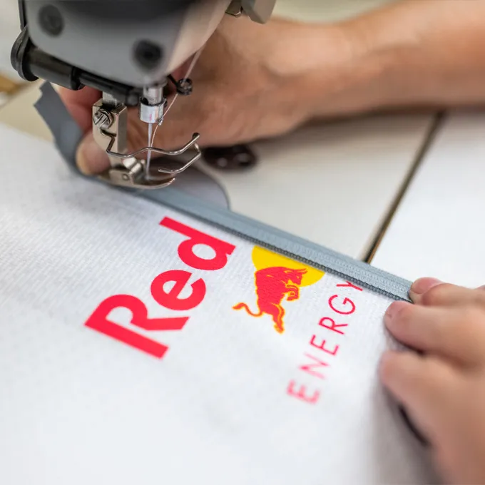 Et nærbilde av en syemaskin som syr en hvit sportstøy med Red Bull-logoen, mens en person holder stoffet.