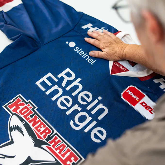 En person sjekker en blå tilpasset sportskjorte fra owayo med ulike sponsorlogoer, inkludert 'Rhein Energie' og 'Kölner Haie'.
