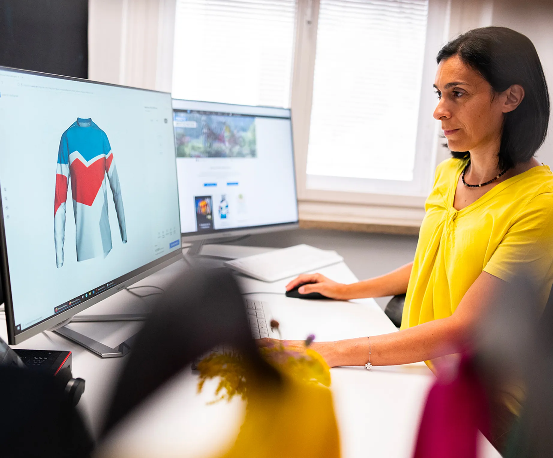 En kvinne i en gul topp sitter ved et skrivebord og designer en tilpasset sportskjole med blå, røde og hvite paneler på en dataskjerm.