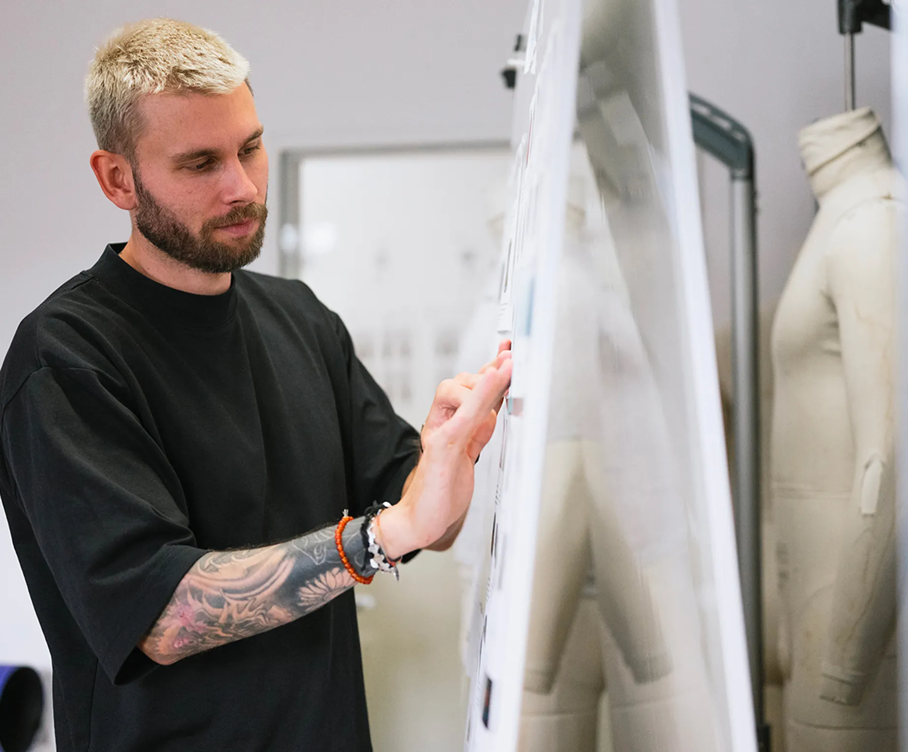 En mann med blondt hår og et tatovering på armen ser konsertrert på design på en klemmbrett i et designstudio, mens en skjørtmannikin står i bakgrunnen.